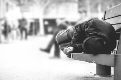 Poor homeless man or refugee sleeping on the wooden bench on the urban street in the city, social documentary concept, selective focus, black and white